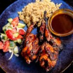 Two bbq quail breasts and thigh on a place with a cup of bourbon bbq sauce and a bed of rice and salad.