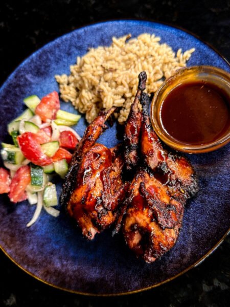 Two bbq quail breasts and thigh on a place with a cup of bourbon bbq sauce and a bed of rice and salad.