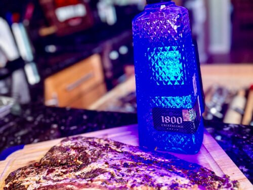 iberico pork on a wooden cutting board next to a bottle of blue illuminated tequila.