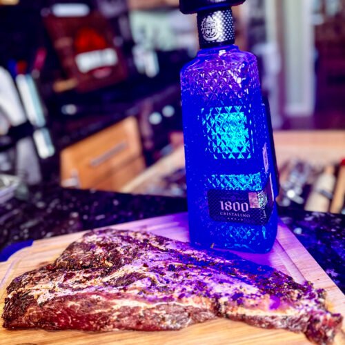 iberico pork on a wooden cutting board next to a bottle of blue illuminated tequila.
